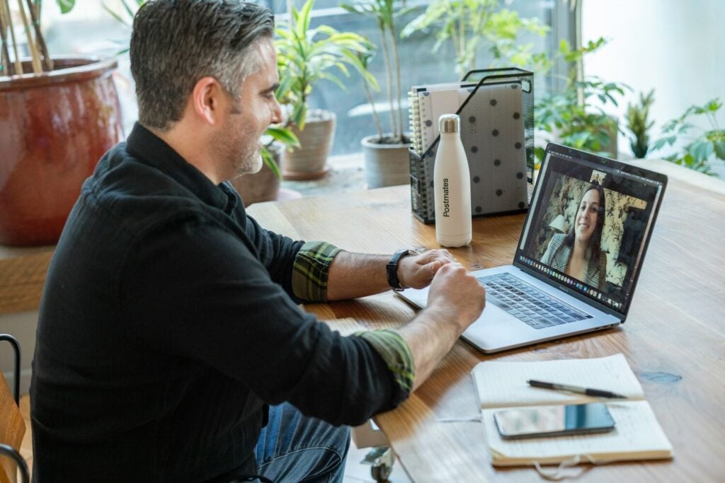 Man sitting at a wooden desk having a video call on a laptop in a bright home office with plants